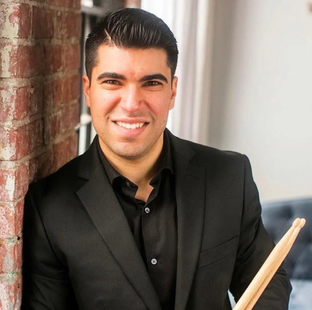 A young man in a black suit smiling holding drumsticks, standing next to a brick wall.